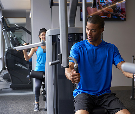 Man in blue shirt using fitness machine with woman in light blue shirt behind him