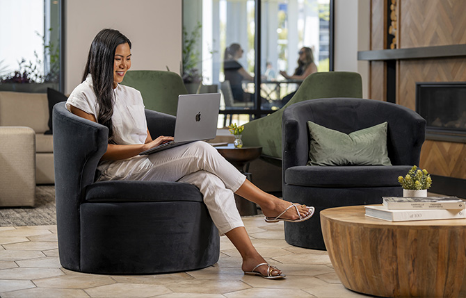 A woman sitting in an armchair with her laptop on her lap working at an AVE lobby 