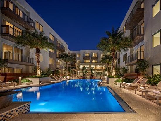 Dusk pool view with lights, pool lounge chairs, palm trees, and views of community in background