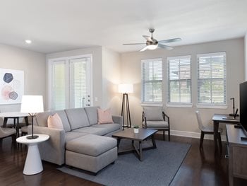 Fully furnished living room with gray sofa, dark wood tables and kitchen in background